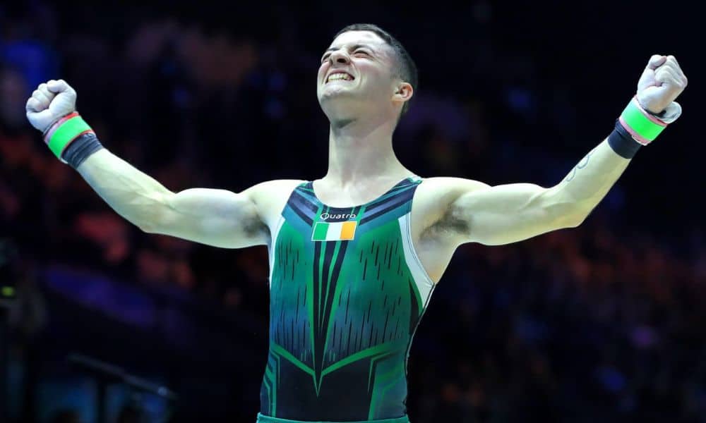 Rhys ergue os braços e sorri. Ele veste uma malha verde com a bandeira da Irlanda no centro