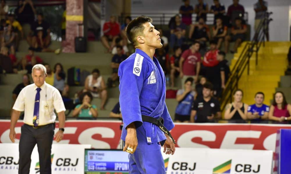 Eduardo Yudy, de pé no dojo, vestindo um quimono azul. No fundo, um árbitro