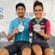 Na imagem, Karen e Rodrigo sentados no lugar mais alto do pódio mostrando as medalhas de vencedores.