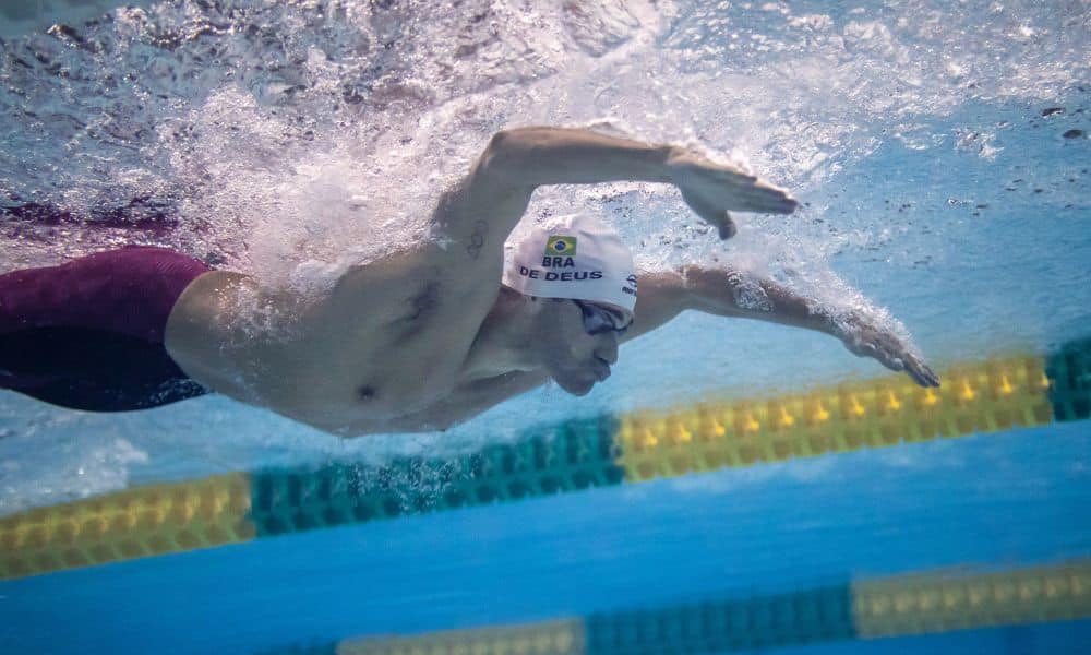 Leonardo de Deus nada em uma piscina. Ele veste uma bermuda púrpura e touca branca e faz o movimento do nado borboleta