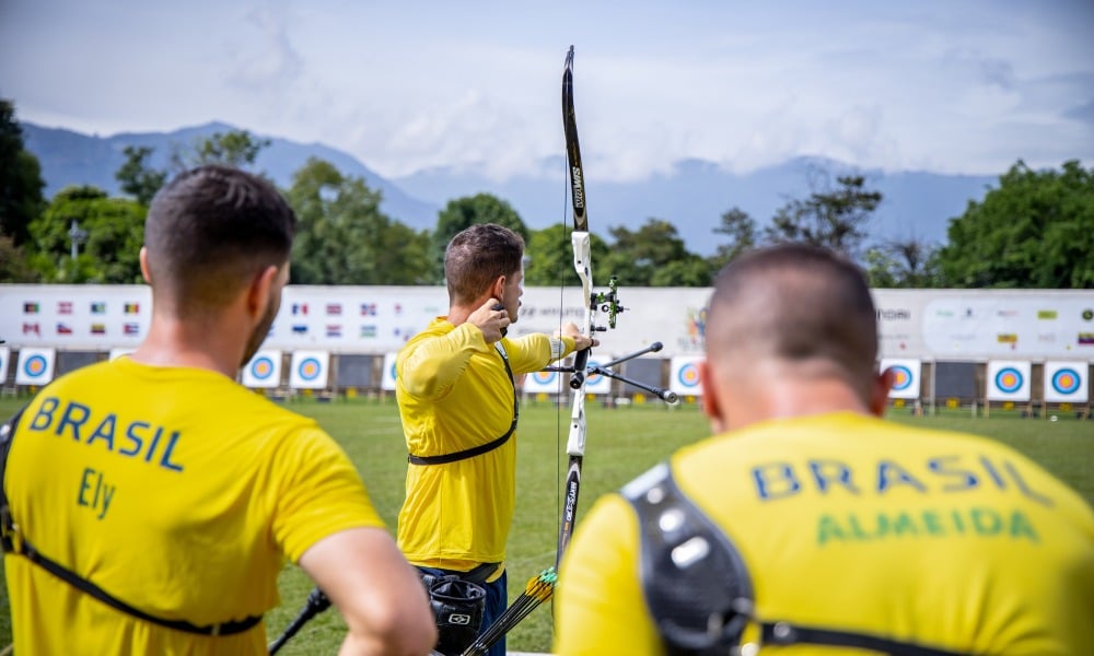 Marcus D'Almeida dispara flecha ao fundo enquanto Matheus Ely e Matheus Almeida observam durante a disputa por equipes do recurvo masculino