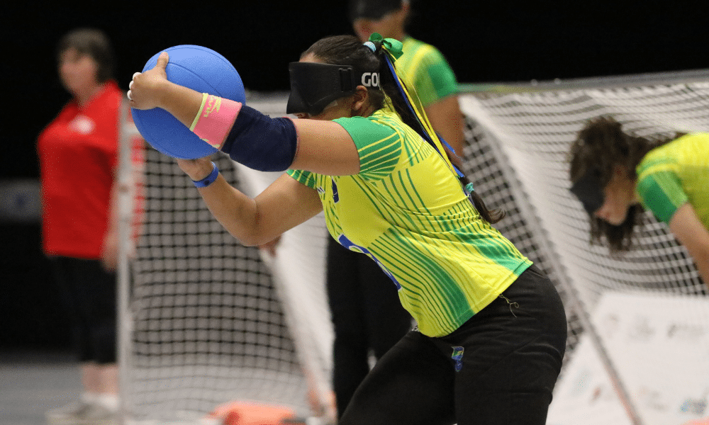 Jogadora do Brasil se preprara para fazer arremesso no goalball. Ela veste uma camiseta amarela e segura a bola azul na quadra dos Jogos Mundiais da IBSA