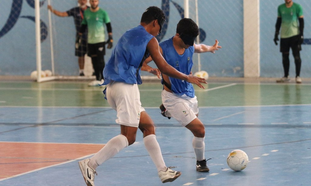 Raynã e Lucas Caetano, que vestem coletes azuis sem camisetas por baixo e calções e meiões azuis, disputam jogada durante treino em uma quadra coberta. Ao fundo, desfocados, aparecem os goleiros Matheus e Giovane. Foto: Renan Cacioli/ CBDV.