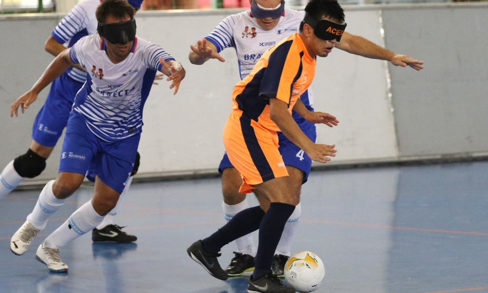 Vestindo uniforme laranja com detalhes em azul marinho, Angel conduz a bola enquanto passa pela marcação de três atletas do INV em partida do Regional Sul-Sudeste de futebol de cegos. Foto: Renan Cacioli/ CBDV.