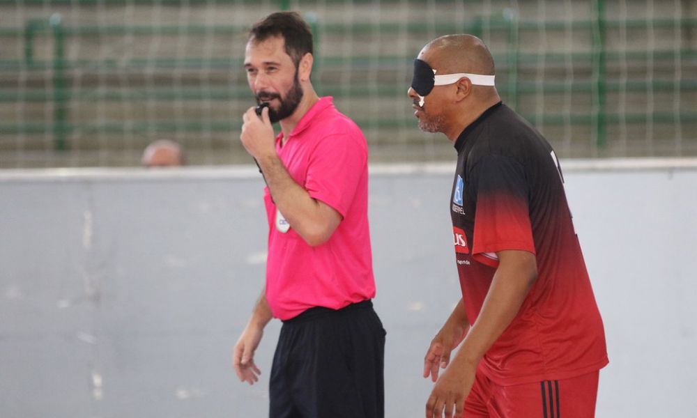 À direita da imagem e usando uma venda preta, o atleta Caio Silva, que é preto e tem o cabelo raspado, espera sinalização do árbitro Raphael Ferreira, que sorri enquanto leva o apito à boca em partida o Regional Sul-Sudeste de futebol de cegos (Foto: Renan Cacioli/ CBDV)