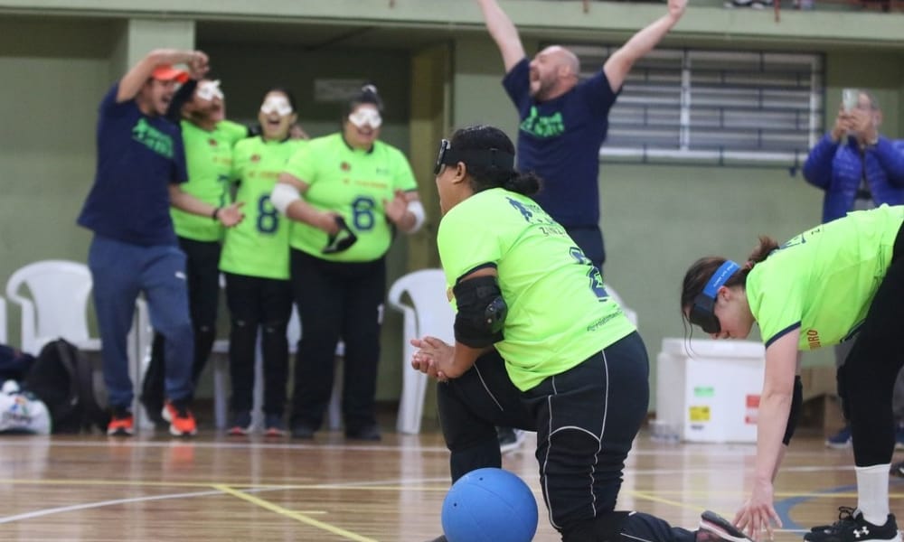 Jimena ergue o braço direito segurando a bola azul para tomar o impulso do arremesso. A foto foi tirada de trás do gol do Maestro.