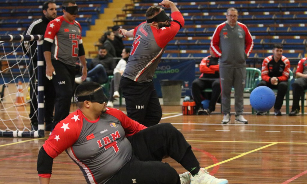 Time masculino do IRM no Regional Sul de Goalball. Um dos atletas está deitado no chão em posição de defesa, enquanto o ouro está de pé após lançar a bola