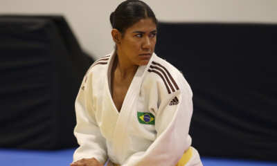 A judoca Kelly Kethyllin, que tem a pela bem morena e o cabelo preto preso em um rabo de cavalo, está apoiando o peso do corpo no joelho direito durante aquecimento, enquanto olha com expressão séria ao lado oposto. Ela veste quimono branco.