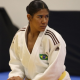 A judoca Kelly Kethyllin, que tem a pela bem morena e o cabelo preto preso em um rabo de cavalo, está apoiando o peso do corpo no joelho direito durante aquecimento, enquanto olha com expressão séria ao lado oposto. Ela veste quimono branco.